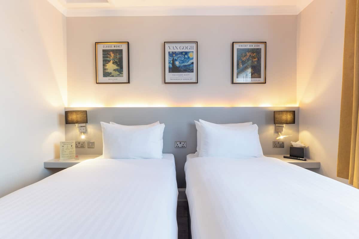 A hotel room with two beds, a headboard, wall-mounted lamps, and three framed artworks above the beds.