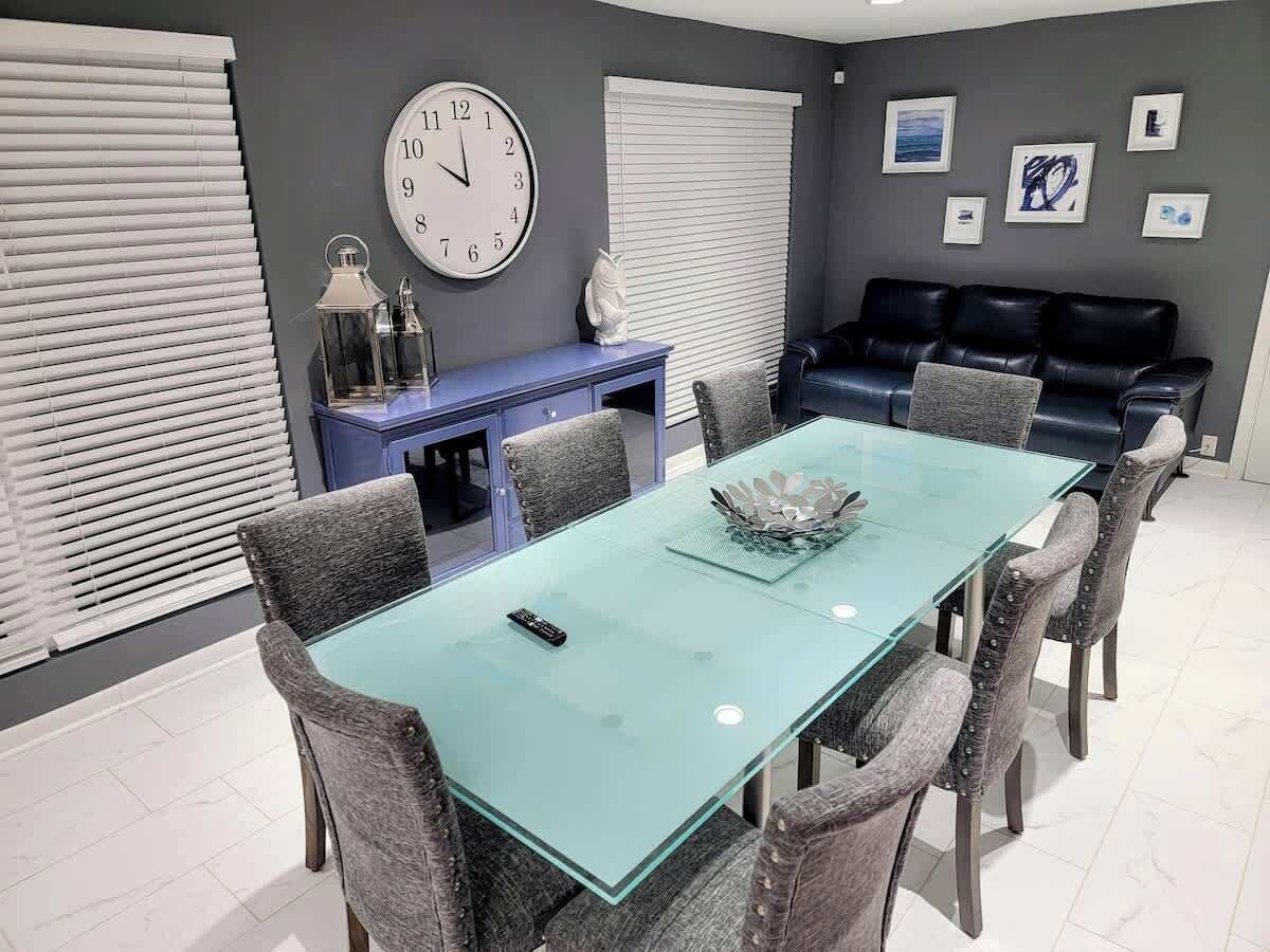 Een eetgedeelte met een glazen tafel, grijs gestoffeerde stoelen, een blauwe console met decoratieve voorwerpen, een zwarte leren bank en een wandklok.