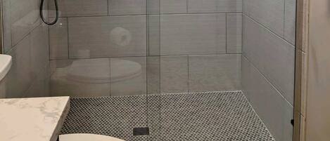 A modern bathroom with a shower, tiled walls, a marble countertop, and a small shelf with toiletries.