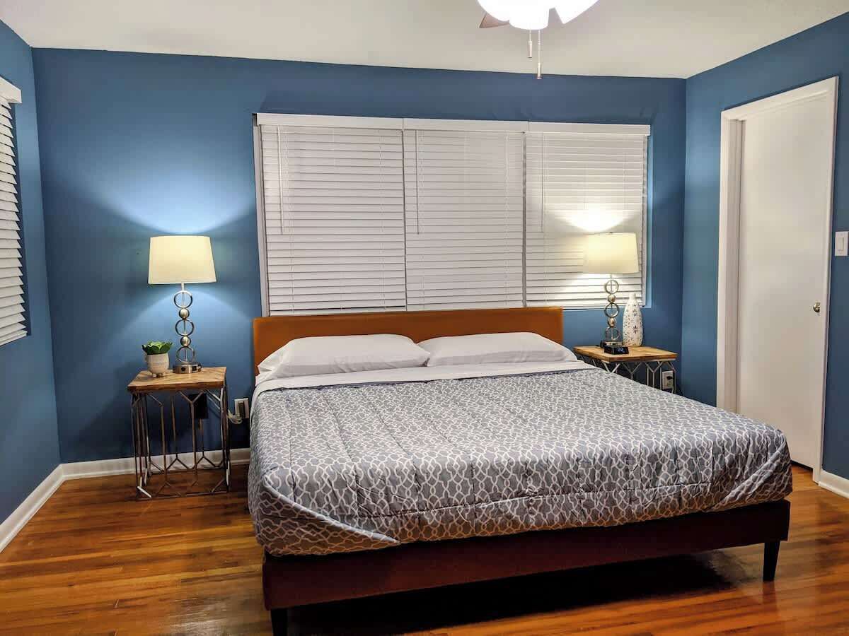 A bedroom with a bed, two bedside tables, a nightstand, a window with blinds, and a door.