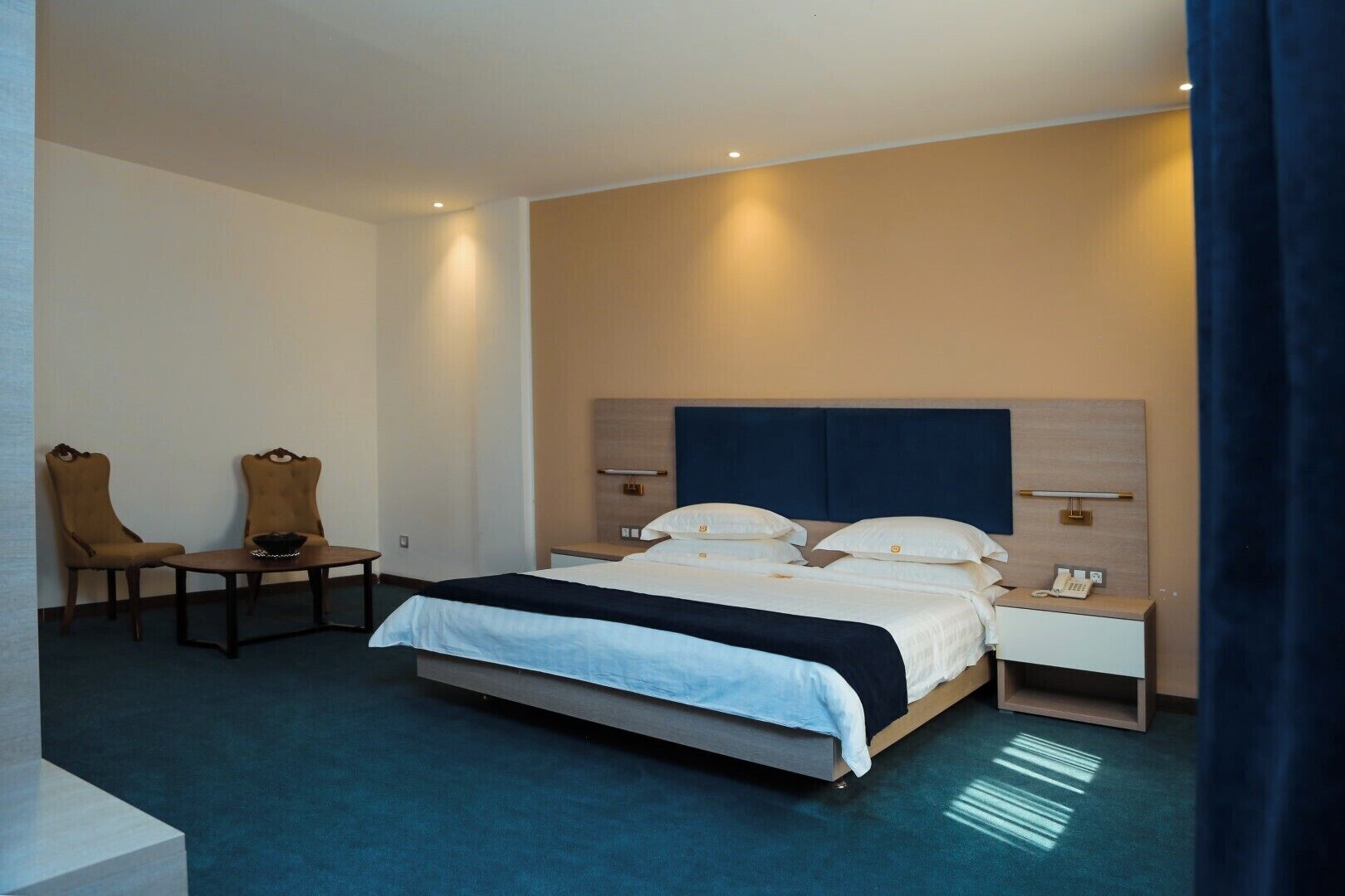 A hotel room with a large bed, two chairs, a small table, a nightstand, and a bedside table.