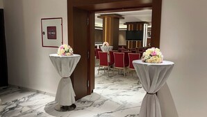A hotel corridor with marble flooring, two round tables with white cloths and floral centerpieces, and a view into a dining area with red chairs and a TV screen. - Al Bayt Hotel (Djelfa)