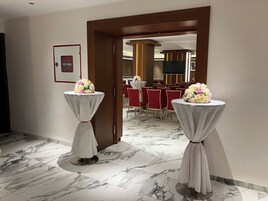 Pasillo de hotel con suelo de mármol, dos mesas redondas cubiertas con manteles blancos y centros de mesa florales, y vista a un comedor con sillas rojas y una pantalla de televisión.