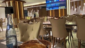 A hotel dining area with a large screen displaying TV channels, comfortable seating, and a bottle of water on the table. - Al Bayt Hotel (Djelfa)