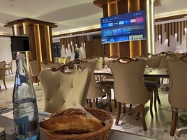 Un comedor de hotel con una pantalla grande mostrando canales de televisión, asientos cómodos y una botella de agua sobre la mesa.