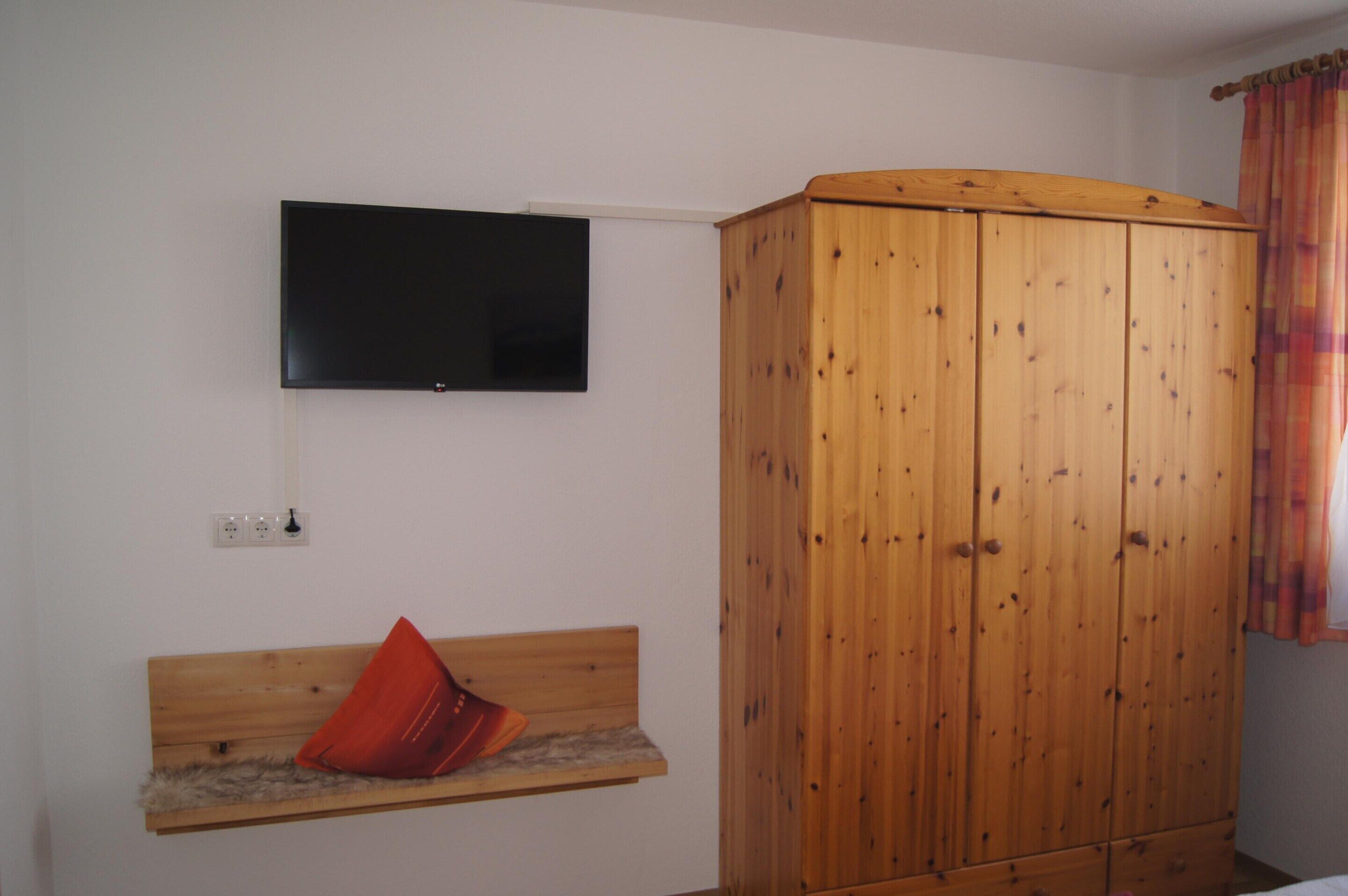 A room with a wooden wardrobe, a mounted flat-screen TV, and a bench with a red cushion.