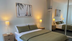 A neatly made bed with a green blanket, two bedside lamps, a wooden nightstand, a small drawer, and a wall-mounted painting of a beach scene.