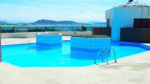 Una piscina en la azotea con agua azul cristalina, dos jacuzzis con mosaicos y vistas a montañas lejanas y una masa de agua.