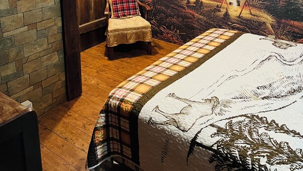 A room with a bed, a plaid chair, a stone wall, and a mural of a forest scene.