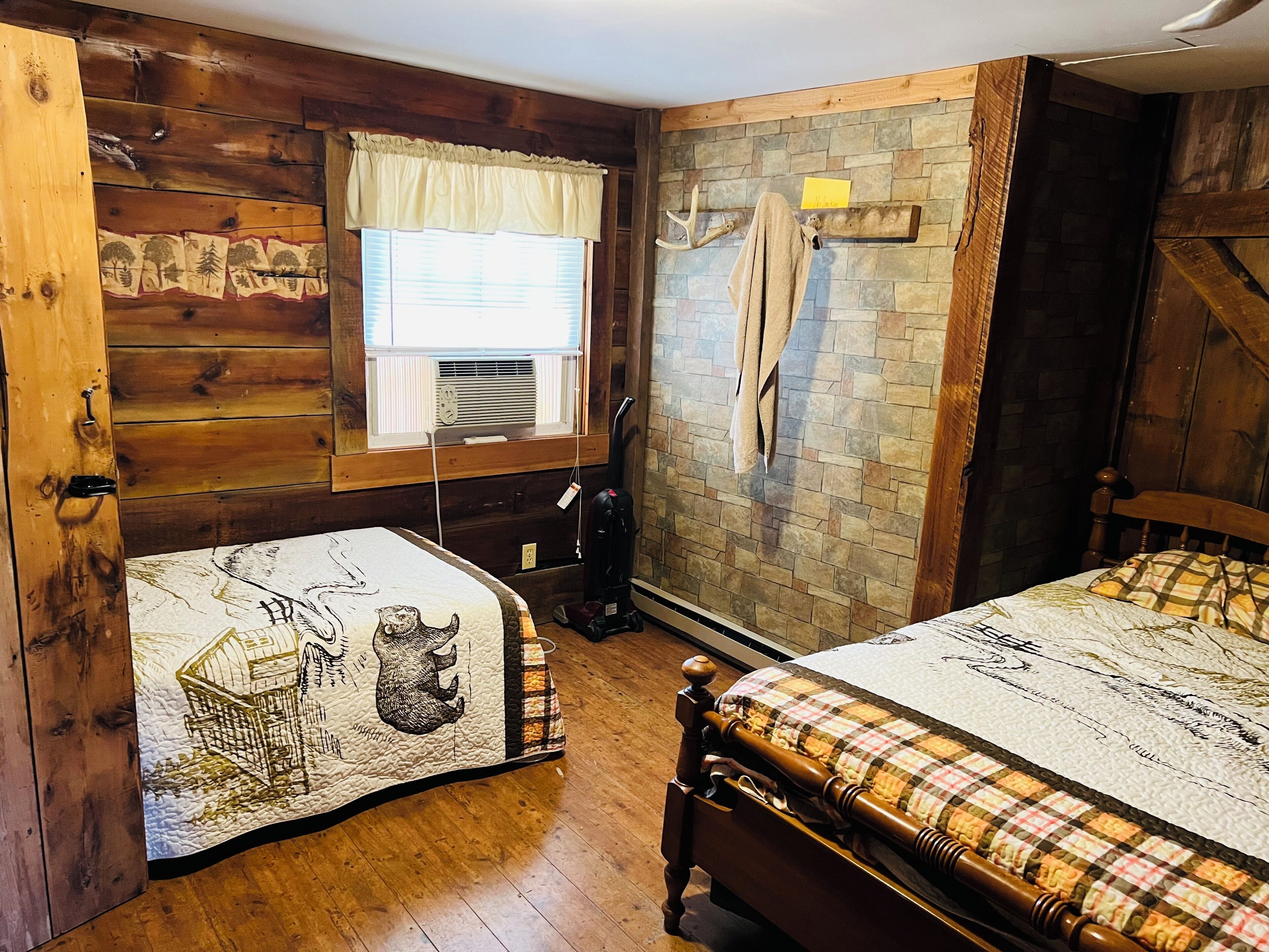 Une pièce avec deux lits, des murs en bois, une fenêtre avec des stores, un mur en pierre orné de divers objets et une porte en bois.