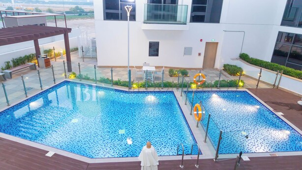 A swimming pool with a tiled deck, surrounded by a building with large windows and a balcony. - Bloomfields 1BR Azizi Gardens Meydan (Dubai)