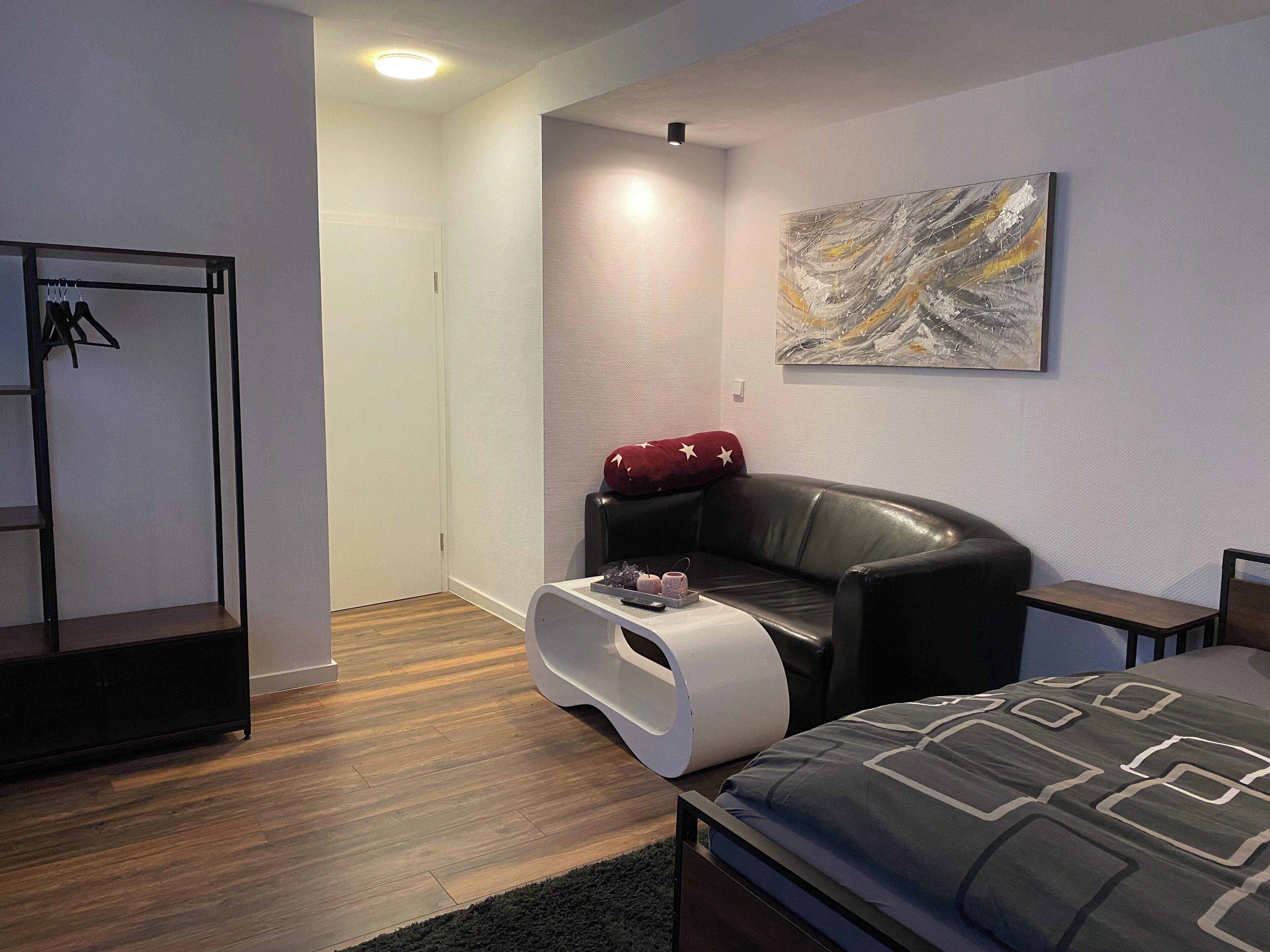 A modern hotel room with a bed, a leather armchair, a small table with a tray, a coat rack, and a painting on the wall.