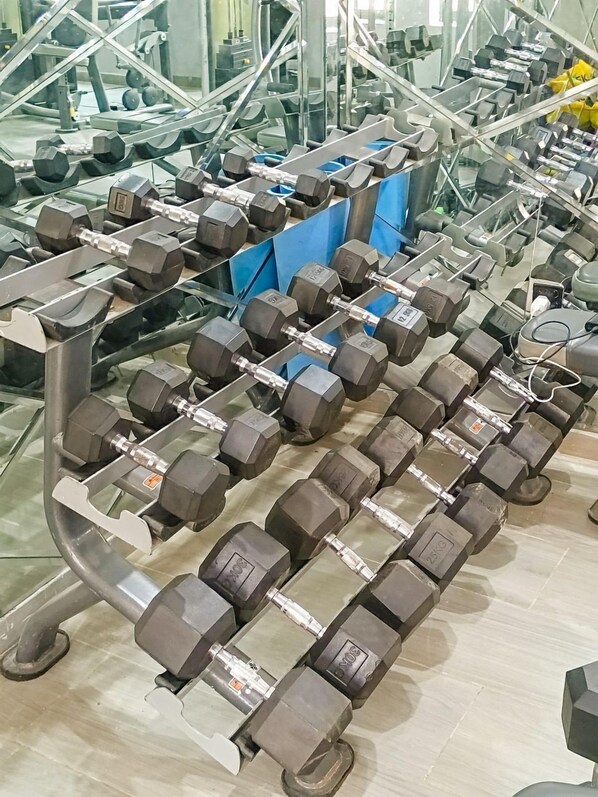 Un gimnasio con filas de mancuernas en estantes, una colchoneta azul y equipo de ejercicio al fondo.