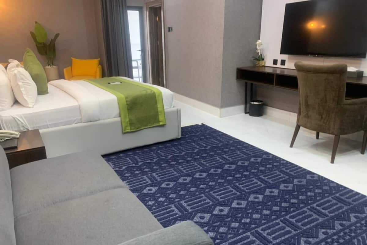 Une chambre d’hôtel moderne avec un lit, un canapé gris, un tapis bleu à motifs, un bureau avec un téléviseur à écran plat et une grande fenêtre.