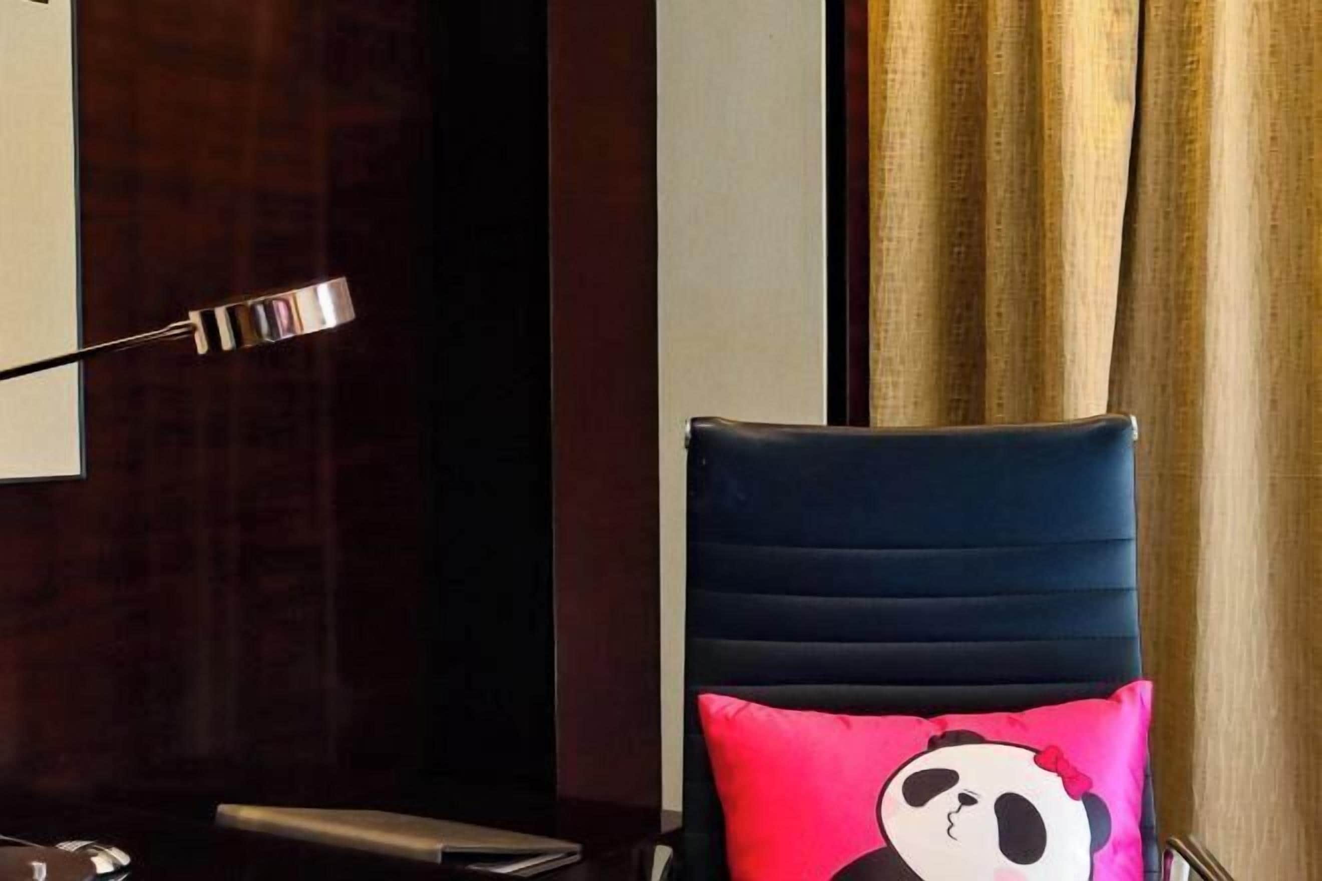 Una habitación con una silla tapizada en azul oscuro, un cojín de panda rojo y blanco, un escritorio con una computadora portátil y una lámpara de pared.