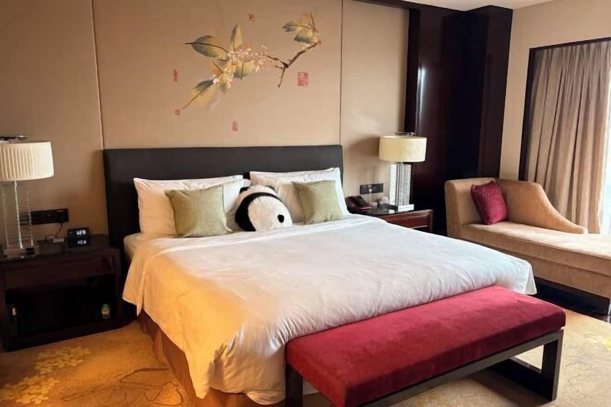 Ein Hotelzimmer mit einem großen Bett, Nachttischen, einem Sessel, einem Wandbild und einem Fenster mit Vorhängen.