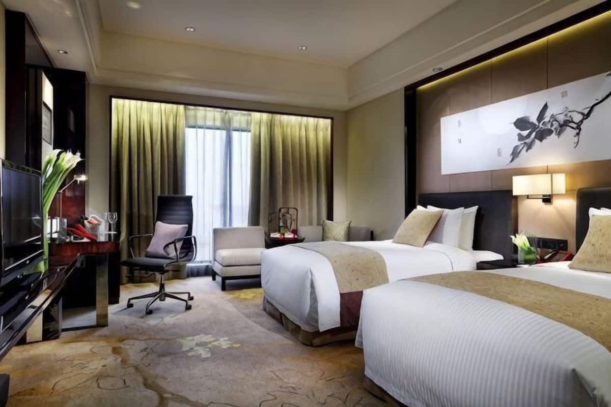 Ein Hotelzimmer mit zwei Betten, einem Schreibtisch mit Fernseher, einem Sessel, einem Fenster mit Vorhängen und einer Wandlampe.