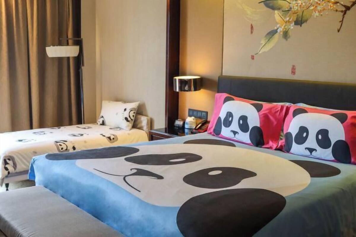 Ein Hotelzimmer mit zwei Betten, jeweils mit Panda-Muster bettbezogen, einem Nachttisch mit Lampe sowie einer Wand mit floralen Bildern.