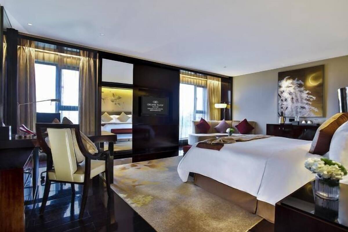 Ein modernes Hotelzimmer mit einem großen Bett, einem Schreibtisch und einem Sessel. Es gibt einen Fernseher, ein Bild an der Wand und ein Fenster mit Vorhängen.
