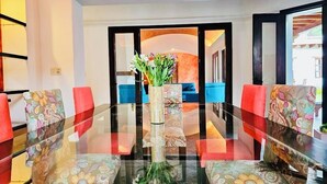 Un comedor con una mesa de vidrio rodeada de sillas coloridas, un centro de mesa floral y vistas a un sofá azul a través de un umbral abierto.