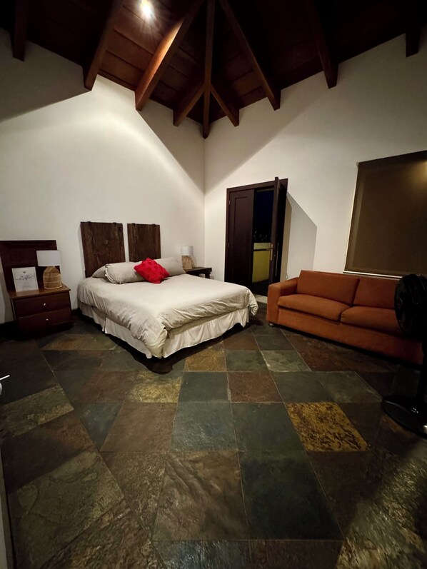 A bedroom with a bed, a wooden headboard, a nightstand, a lamp, a red pillow, a brown sofa, and a large window.