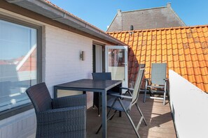 Une terrasse extérieure avec une table et des chaises, une fenêtre et une porte donnant accès à un bâtiment dont le toit est recouvert de tuiles.