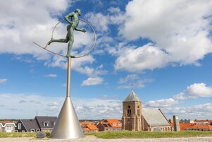 Una estatua de una figura equilibrándose sobre un poste con un aro circular, con una iglesia y edificios residenciales de fondo.
