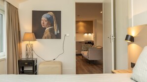 A hotel room with a bed, a nightstand, a lamp, a painting, and an open door leading to a dining area. - Apartment in Zeeland Near the Beach (Zoutelande)