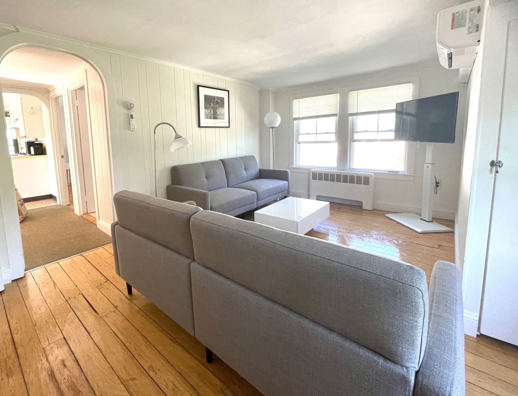 A living room with a grey sofa, a white coffee table, and a TV. There is a door leading to a kitchen and a hallway.