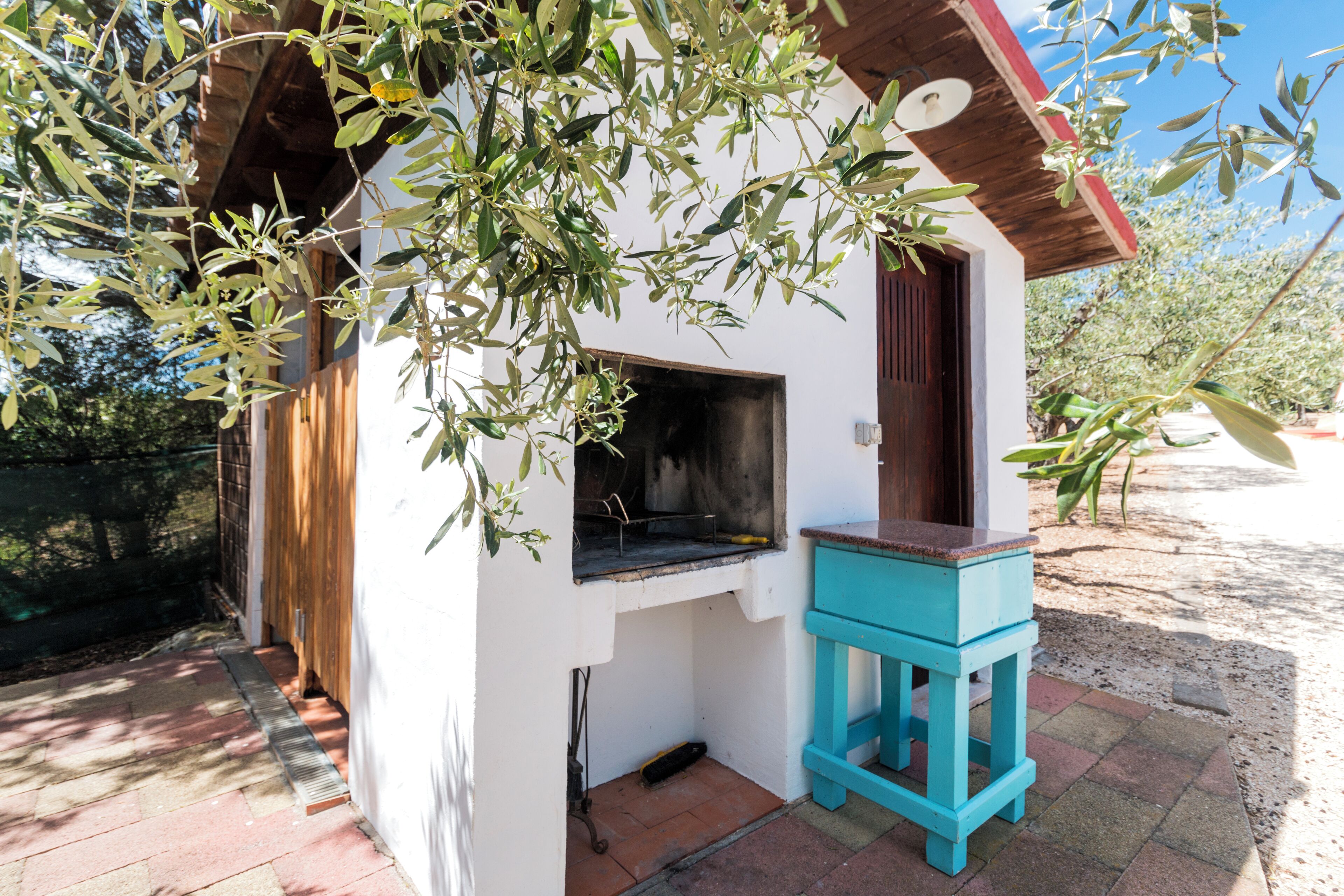 Une petite cuisine extérieure avec un barbecue, un tabouret bleu et un bâtiment blanc doté d’un toit en bois.