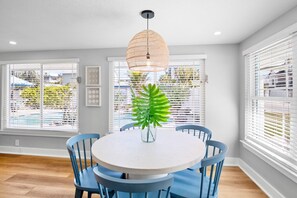 Een eethoek met een ronde tafel, blauwe stoelen, een hanglamp en grote ramen met jaloezieën.