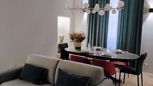 A modern living room with a sofa, dining table, and chairs. There is a chandelier above the table and a vase with dried flowers on it.