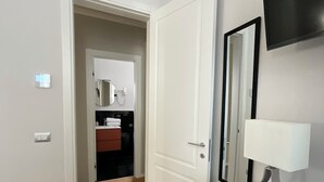 A modern hotel room with a wooden floor, a white dresser, a flat-screen TV mounted on the wall, and a door leading to a bathroom.