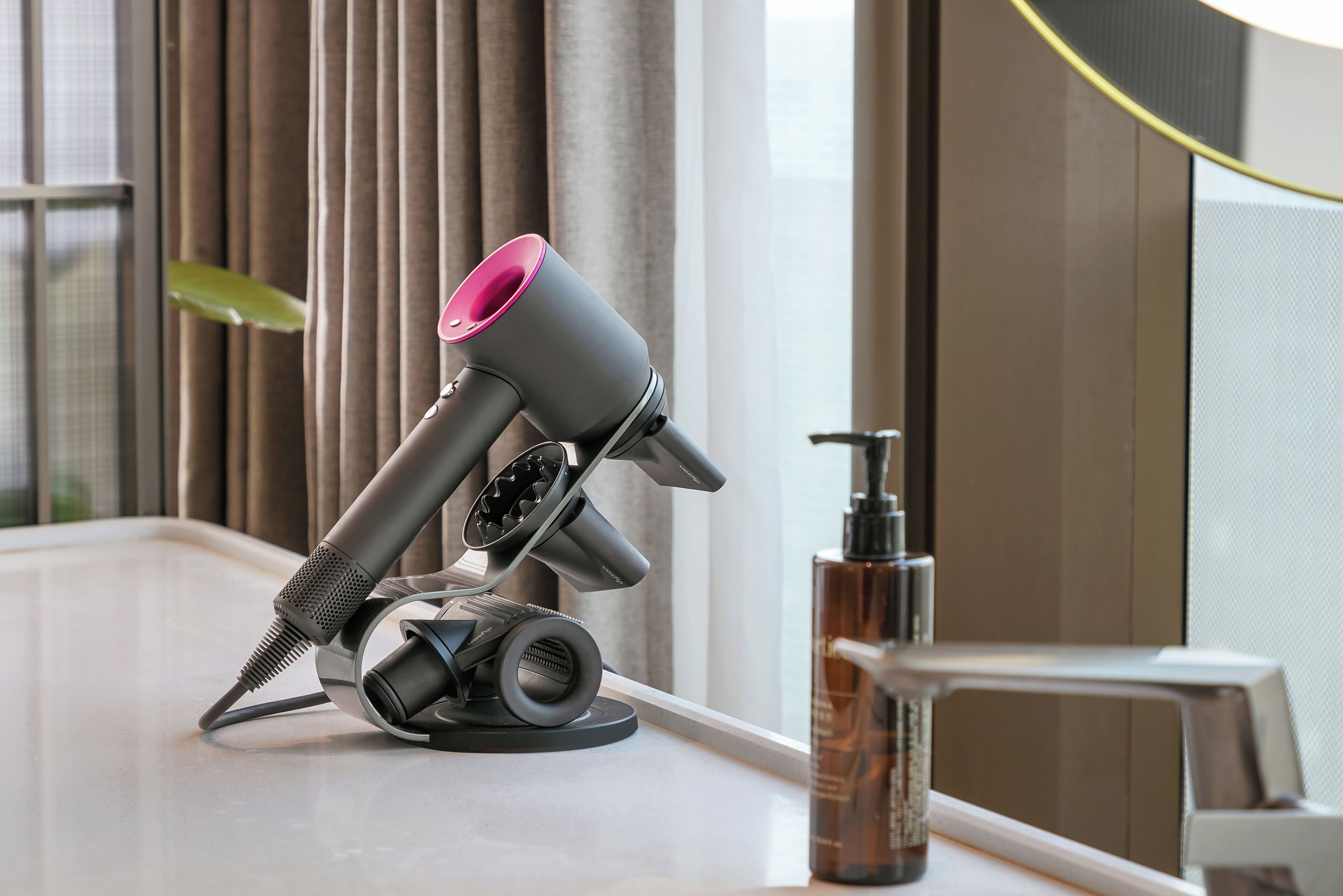 A hair dryer on a counter next to a bottle, with a window and curtains in the background.