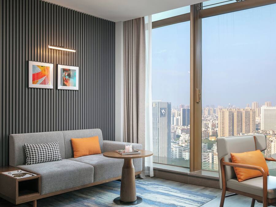 Una habitación de hotel moderna con un sofá gris, una mesita de madera, una mesa de centro redonda y vistas a un paisaje urbano a través de amplios ventanales.