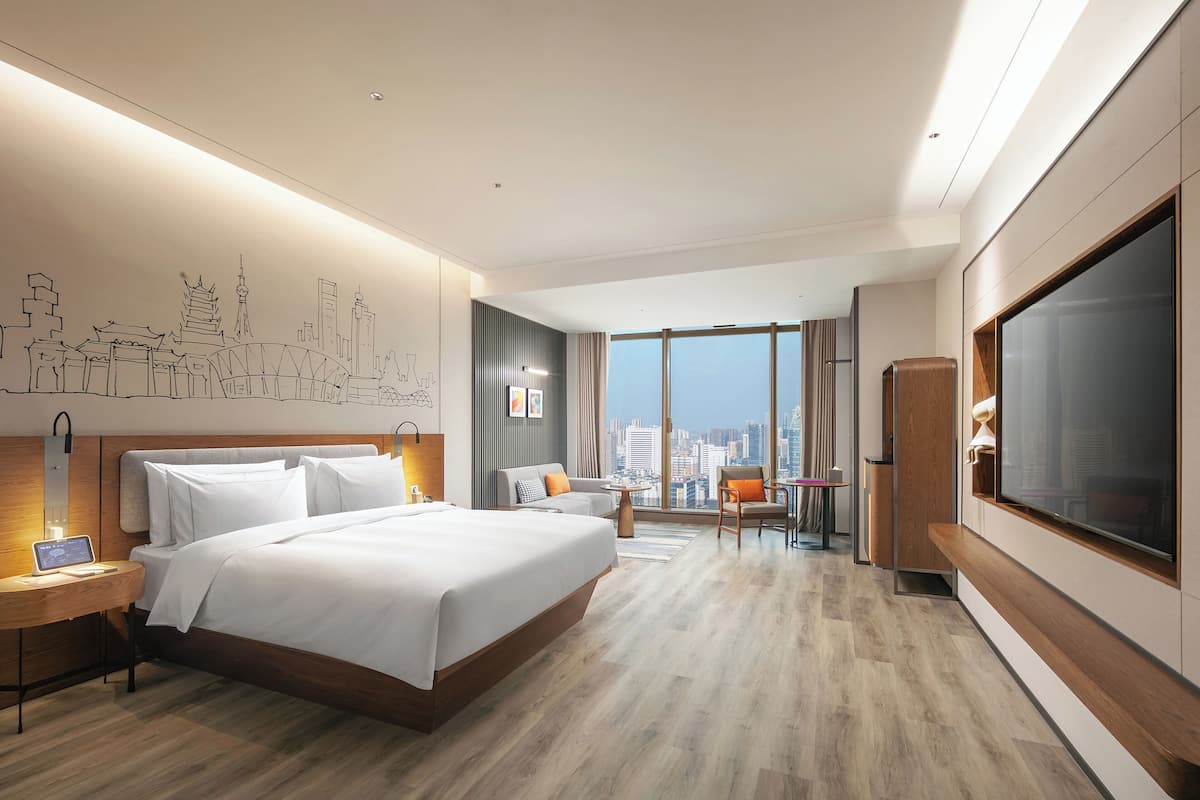 A modern hotel room with a large bed, a desk with a laptop, a TV, and a city view.