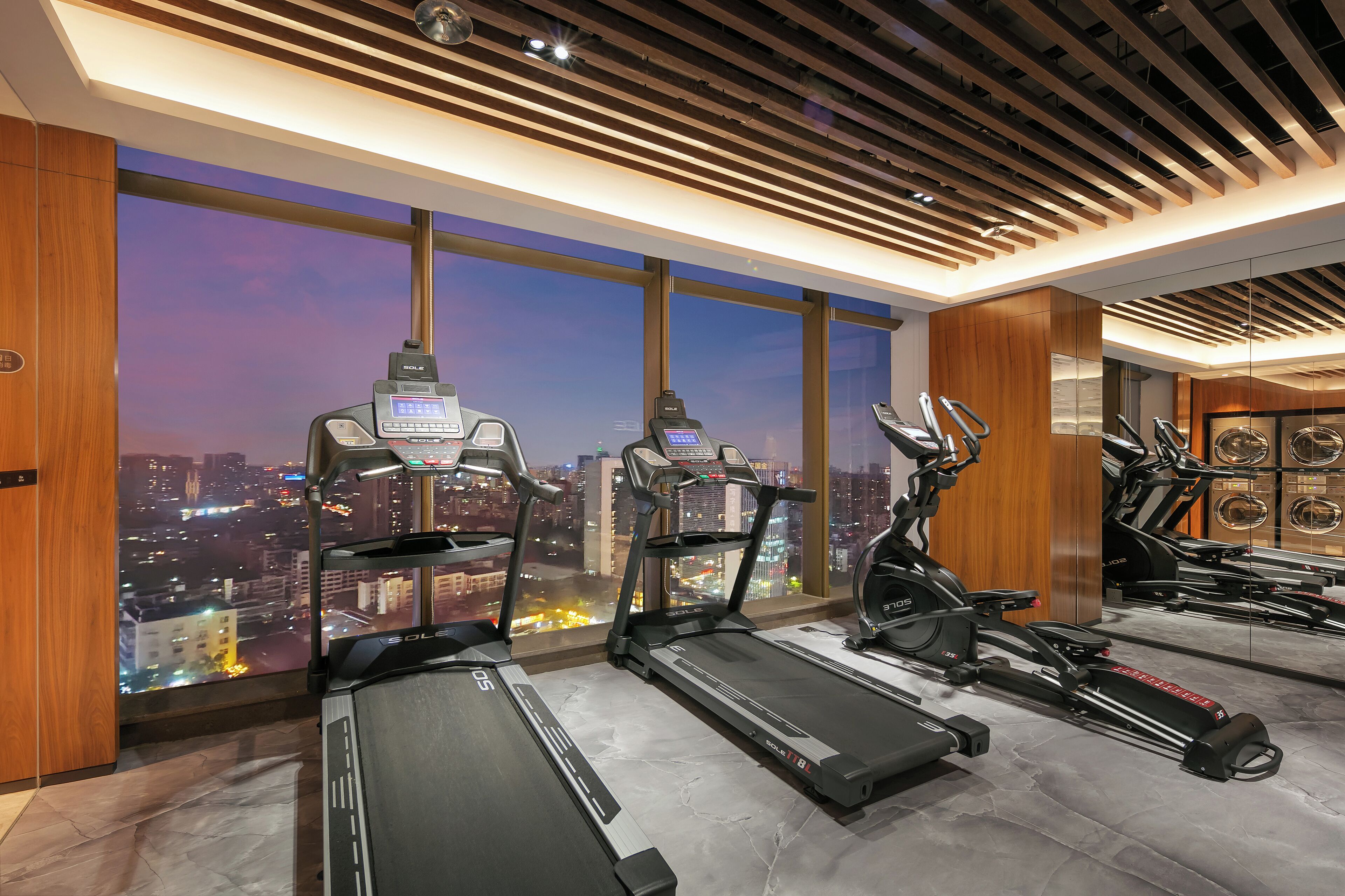 A gym with treadmills, stationary bikes, and a city view through large windows.