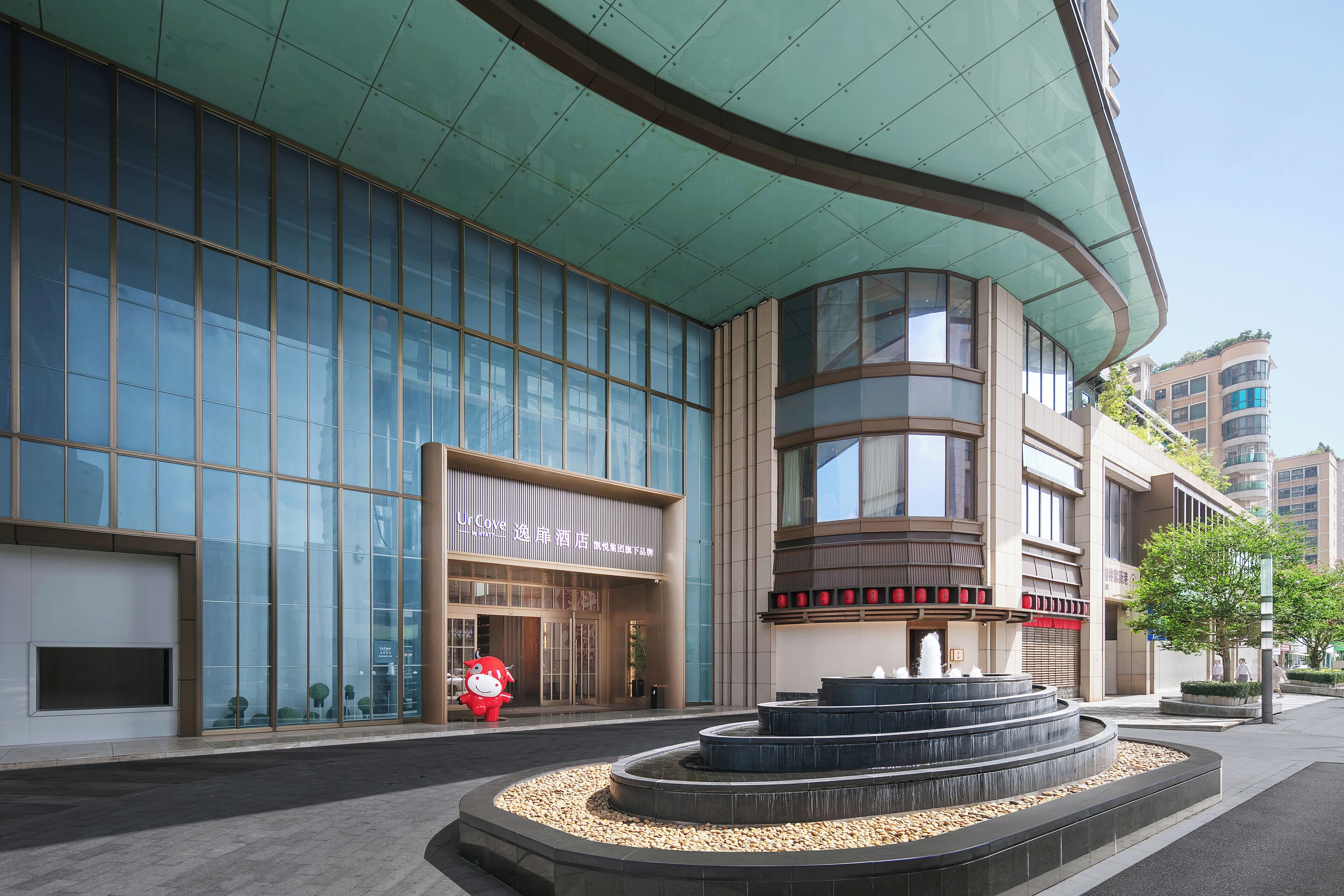 A modern hotel with a curved glass facade, a central fountain, and a red mascot statue outside.