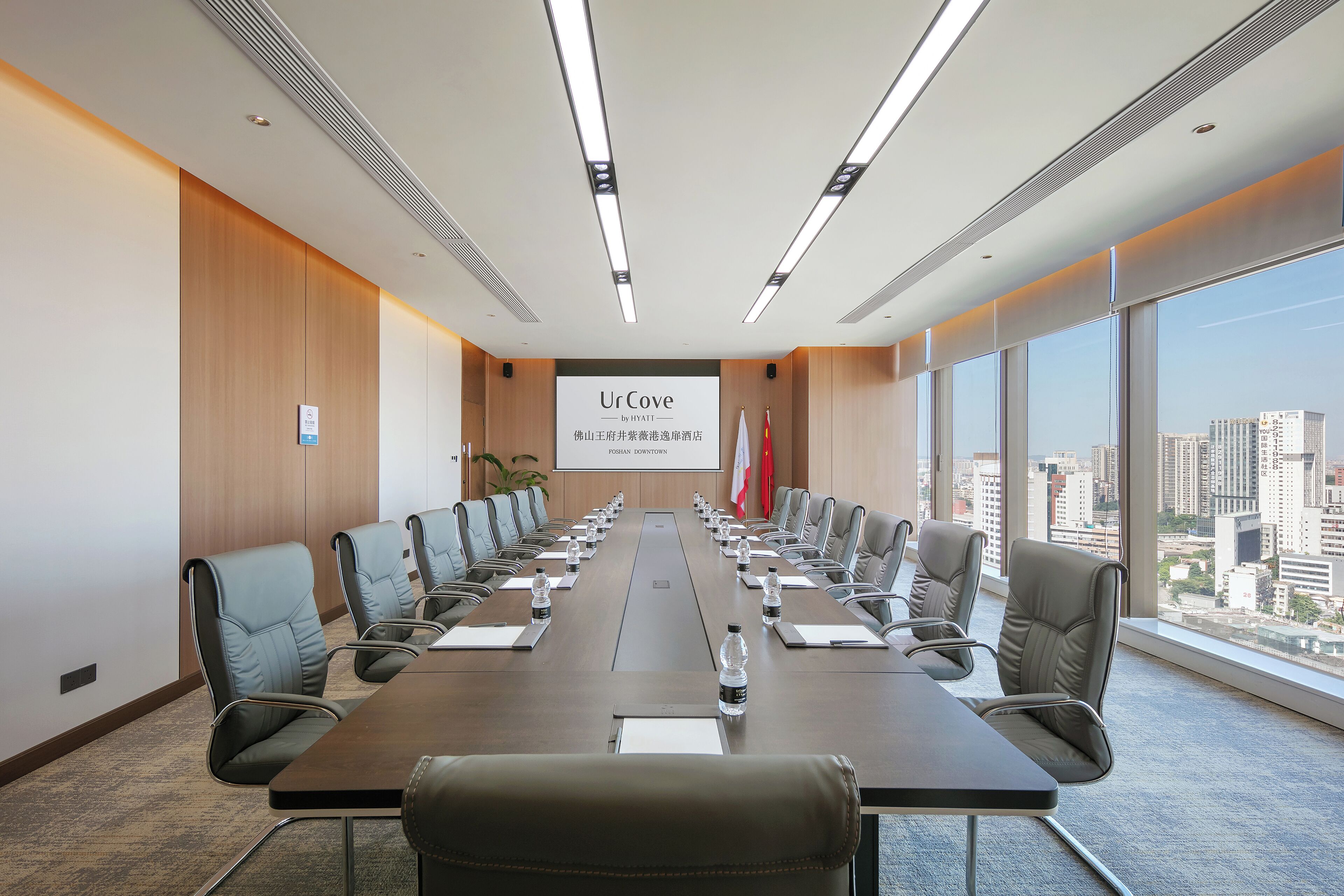 A conference room with a long table, chairs, a projector screen, and a city view through large windows.