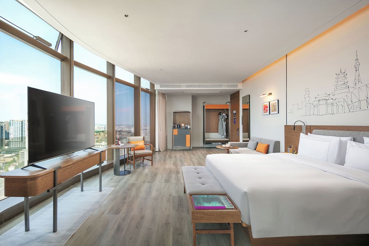 A hotel room with a large TV, a desk, a bed, and a view of the city.