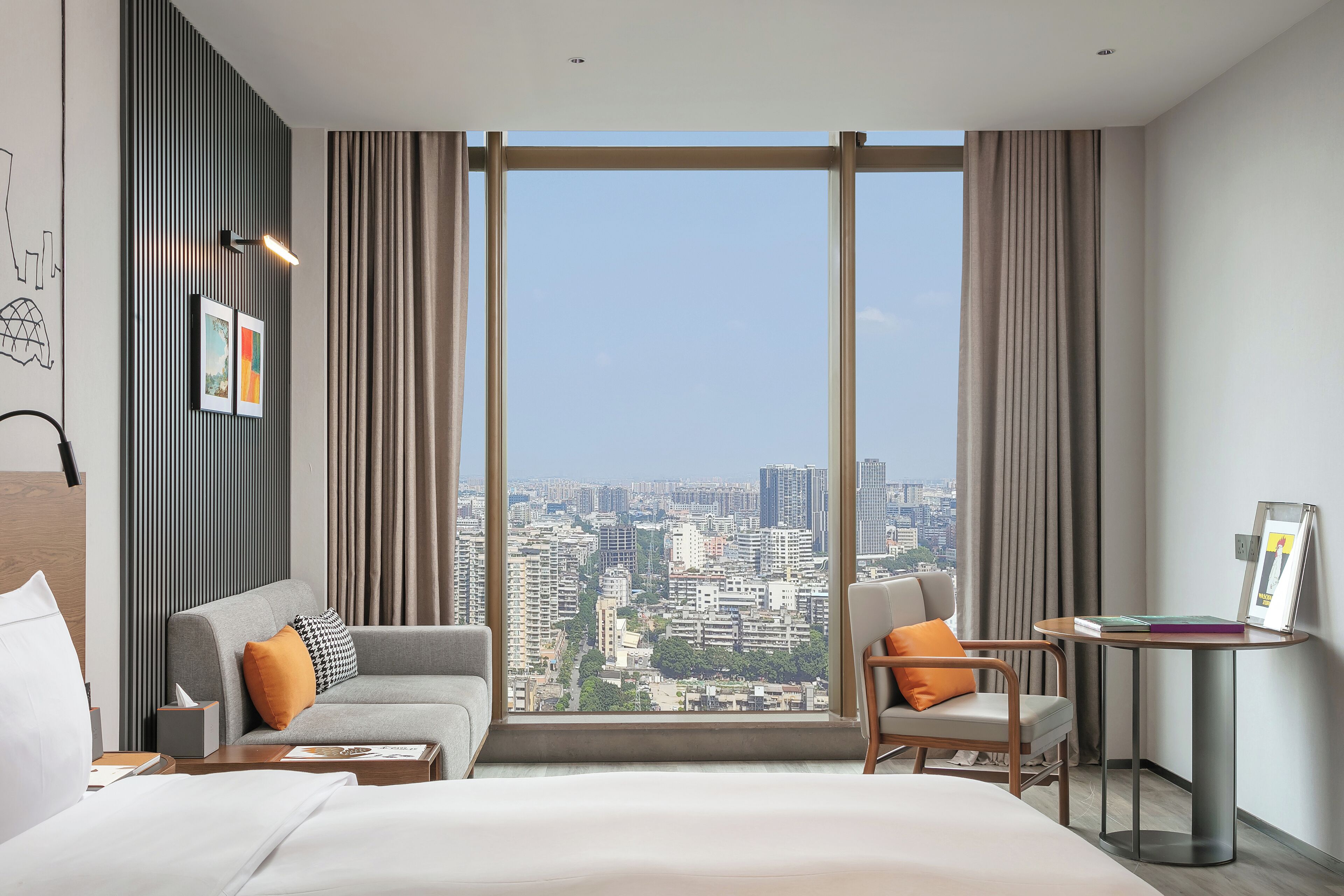 Una habitación de hotel moderna con un ventanal grande, un sofá gris, una silla de madera, una mesita pequeña y una cama.