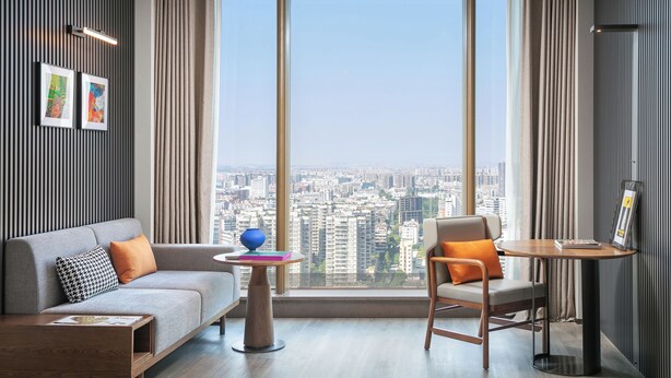 A modern living room with a sofa, a chair, a small table, and a large window offering a city view. - UrCove by Hyatt Foshan Downtown (Foshan)