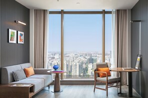 Una sala de estar moderna con un sofá, una silla, una mesita y un amplio ventanal con vistas a la ciudad.