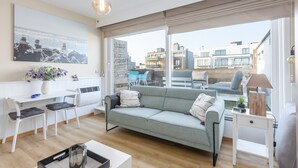 A modern living room with a grey sofa, white chairs, a dining table, and a large window offering a city view. - Penthouse Near Koksijde Beach (Middelkerke)