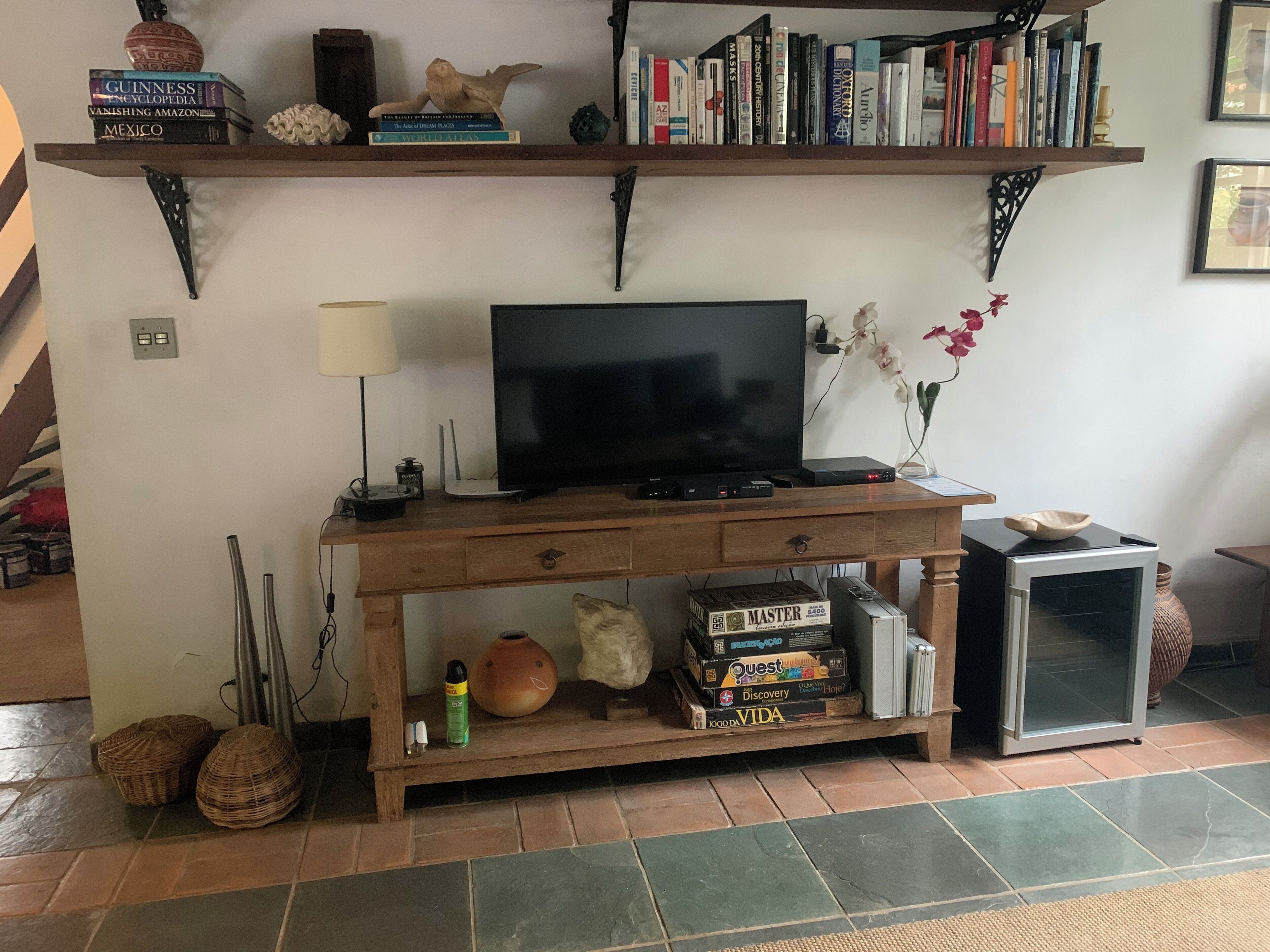 Een woonkamer met een houten tv-meubel, een flatscreen televisie, een boekenkast vol boeken, een bijzettafeltje met een lamp en een decoratieve vaas met bloemen.