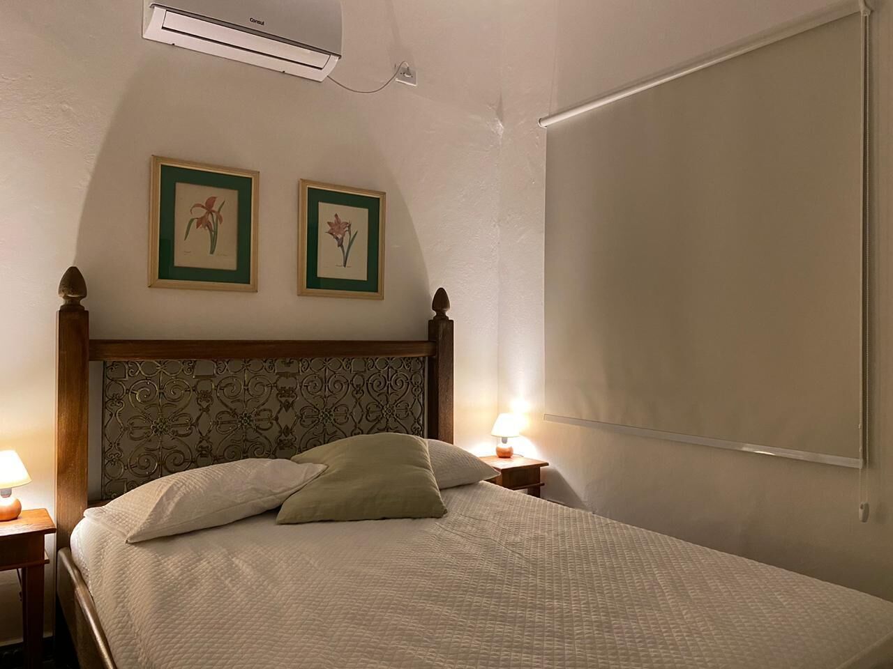 A bedroom with a bed, two framed pictures on the wall, a bedside table with a lamp, and an air conditioning unit.