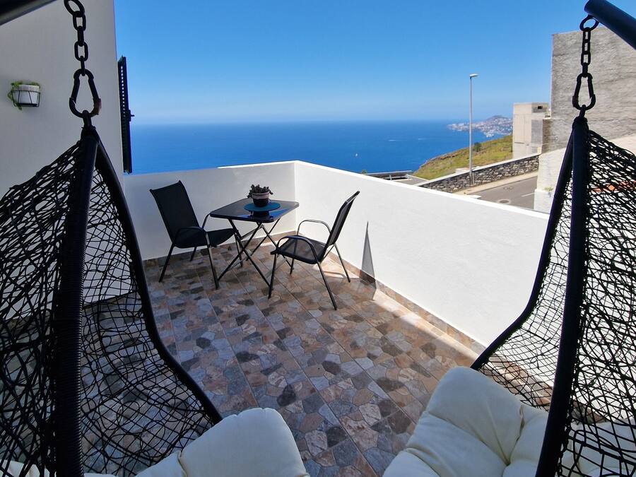 Un balcon avec vue sur l’océan, une table avec des chaises et deux fauteuils suspendus.