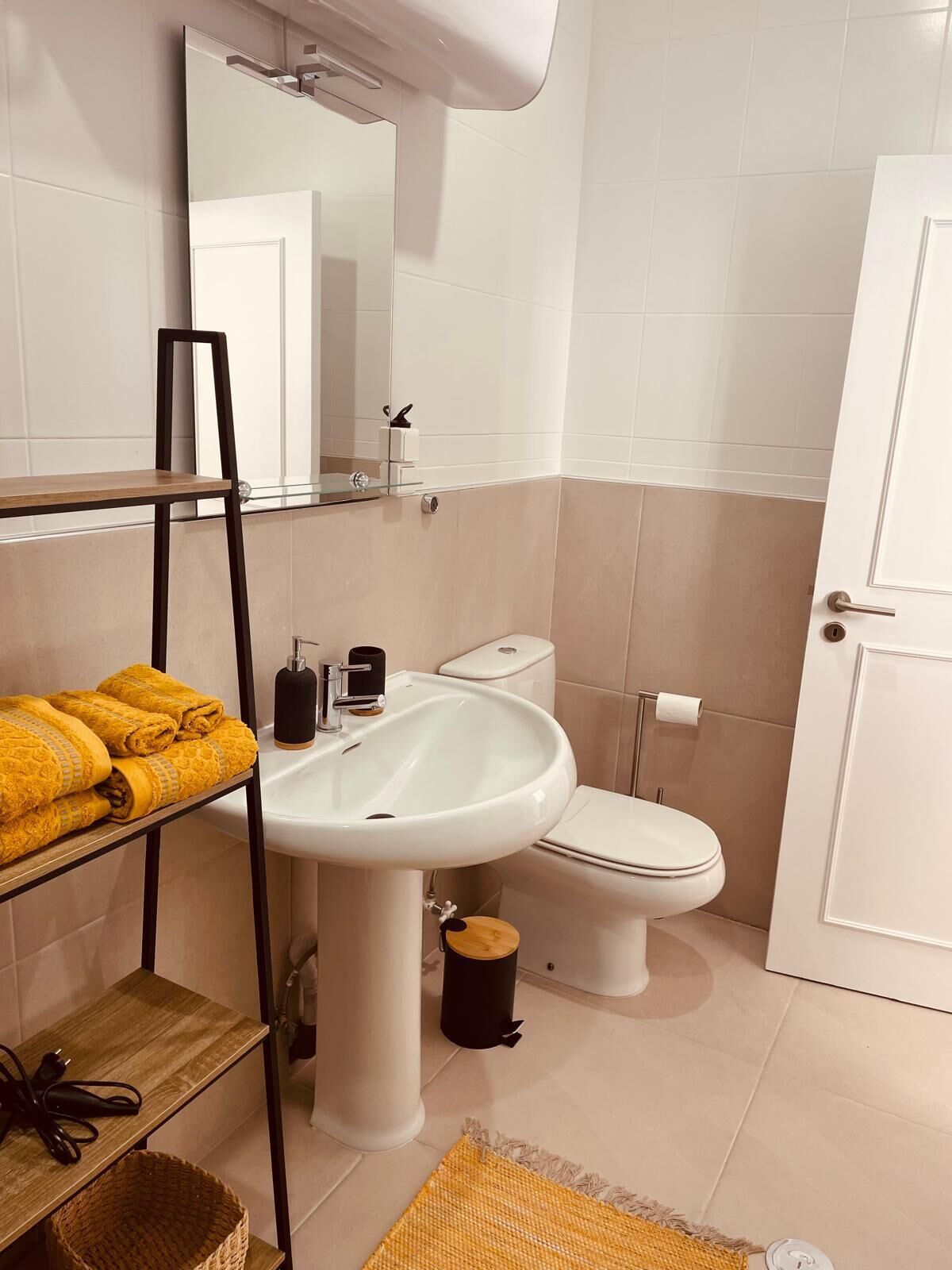 A bathroom with a white sink, toilet, and a mirror above the sink. There is a shelf with towels and a basket on the floor.