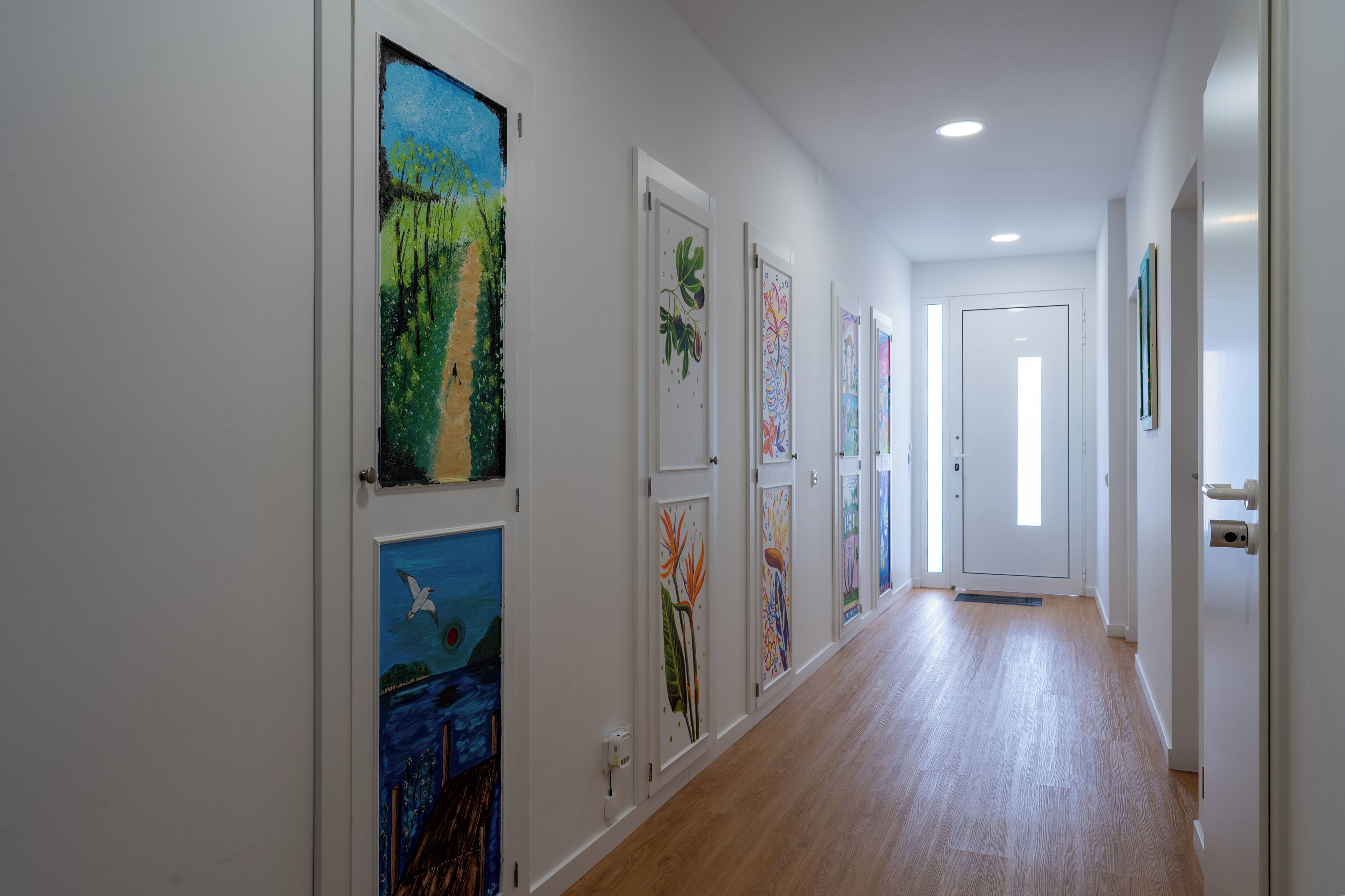 A hallway with wooden flooring and white walls, featuring framed artworks on the left side.