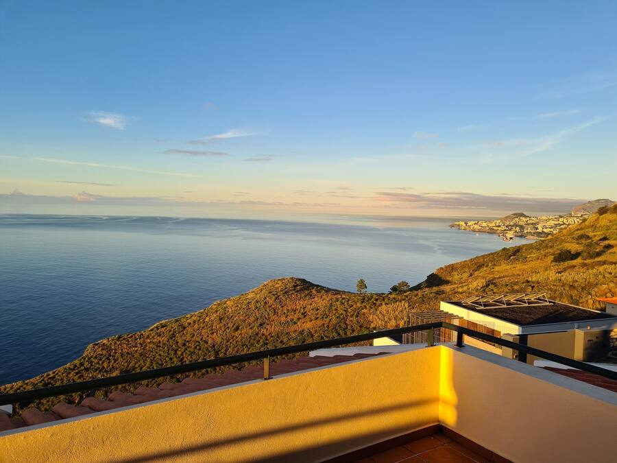 Vistes costaneres des d'un balcó, amb cel serè, mar tranquil·la i un poble a la distància.